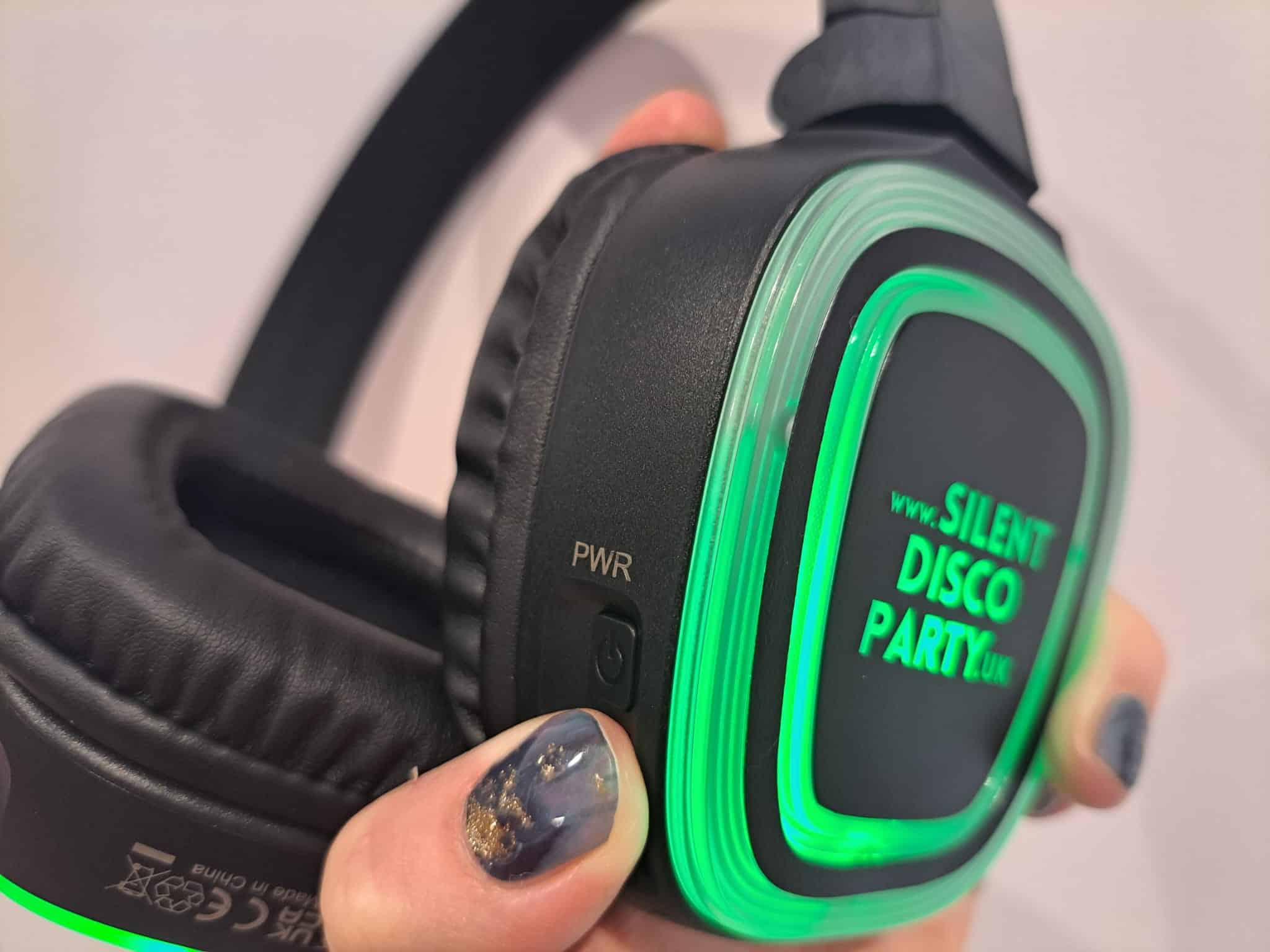 Close up of a Silent Disco Party UK wireless headphone glowing green held in hand showing the branded face panel and PWR power button