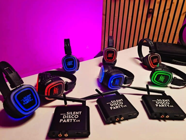 Multiple Silent Disco Party UK wireless headphones glowing red green and blue arranged on a table alongside three branded transmitter boxes against a pink lit background